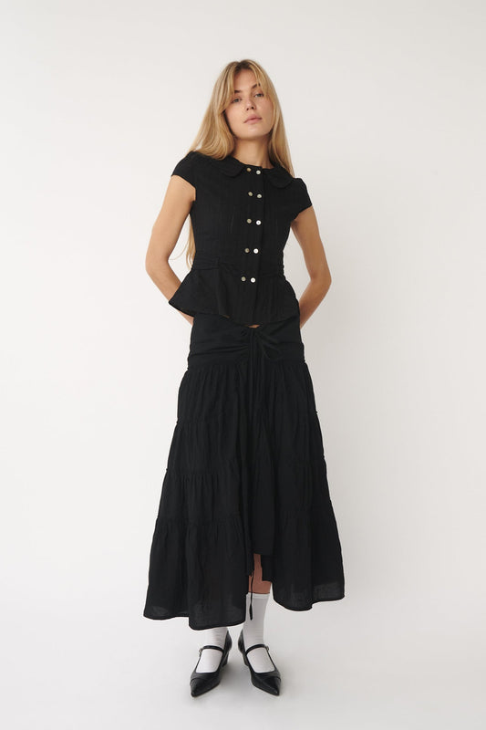 A model wearing a black Virginia Blouse with short sleeves, Peter Pan collar, and eyelet fabric details. The blouse is paired with a long skirt and black shoes.