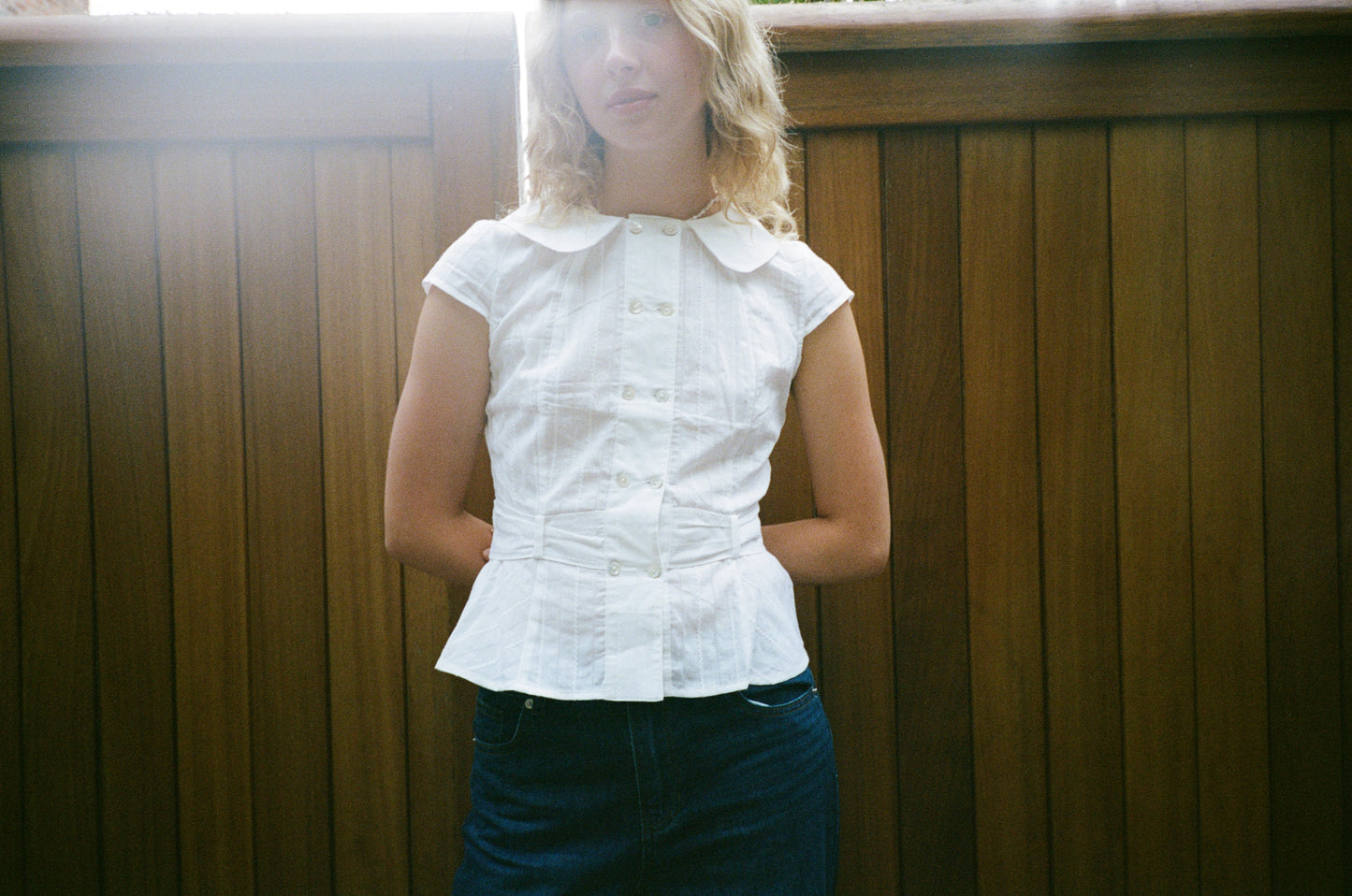 Model wearing the Virginia Blouse cotton in white – a cap-sleeve blouse with double-breasted shell buttons and a Peter Pan collar. In front of wooden fences Independent designer womenswear by A Room London.