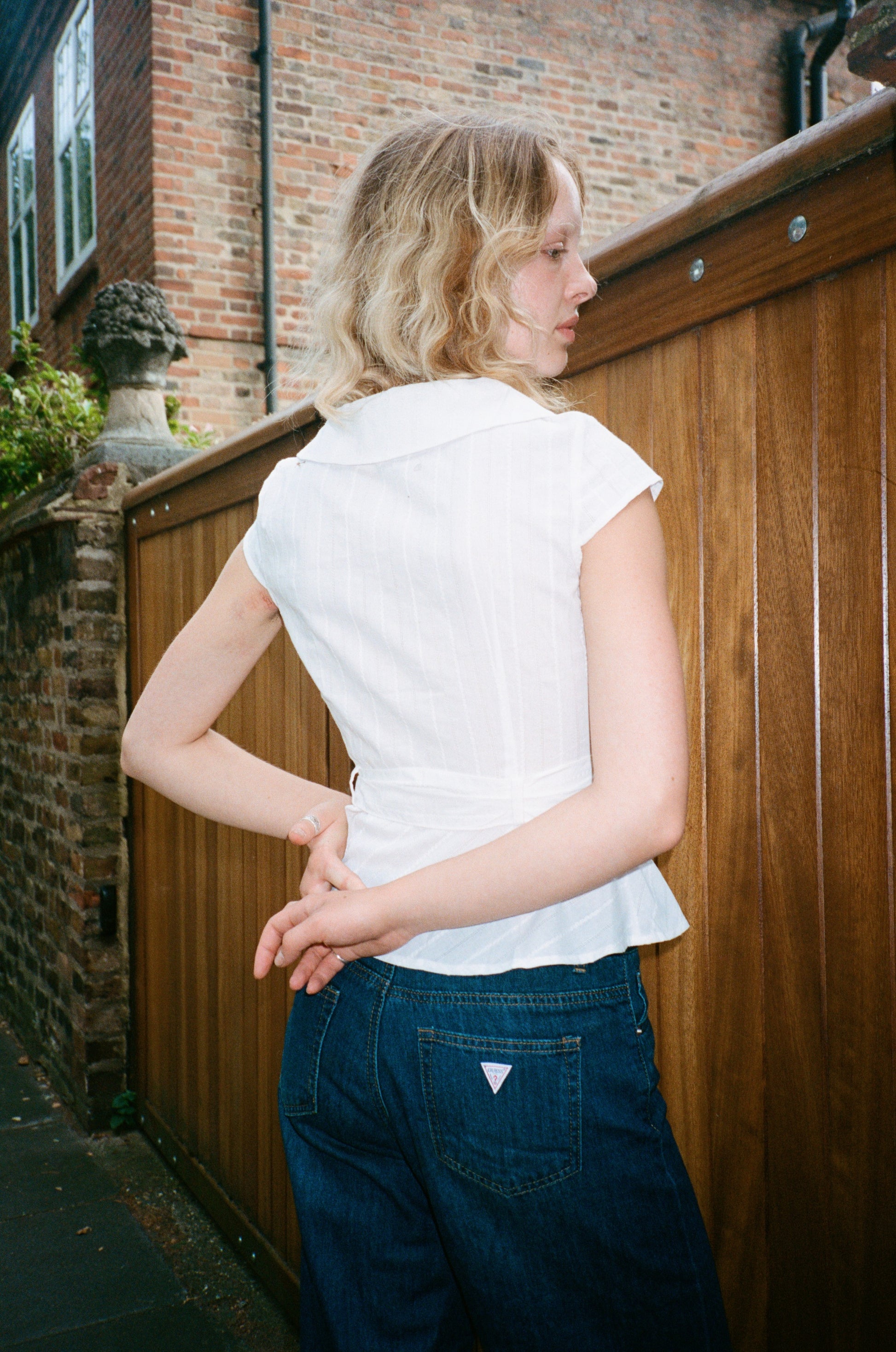 blonde Model wearing the Virginia Blouse cotton in white, back view – a cap-sleeve blouse with double-breasted shell buttons and a Peter Pan collar. Independent designer womenswear by A Room London.
